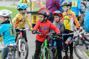 A group of children around 8 years old are on bicycles together, the child in the middle of the picture wears a helmet with a red mohawk down the centre.