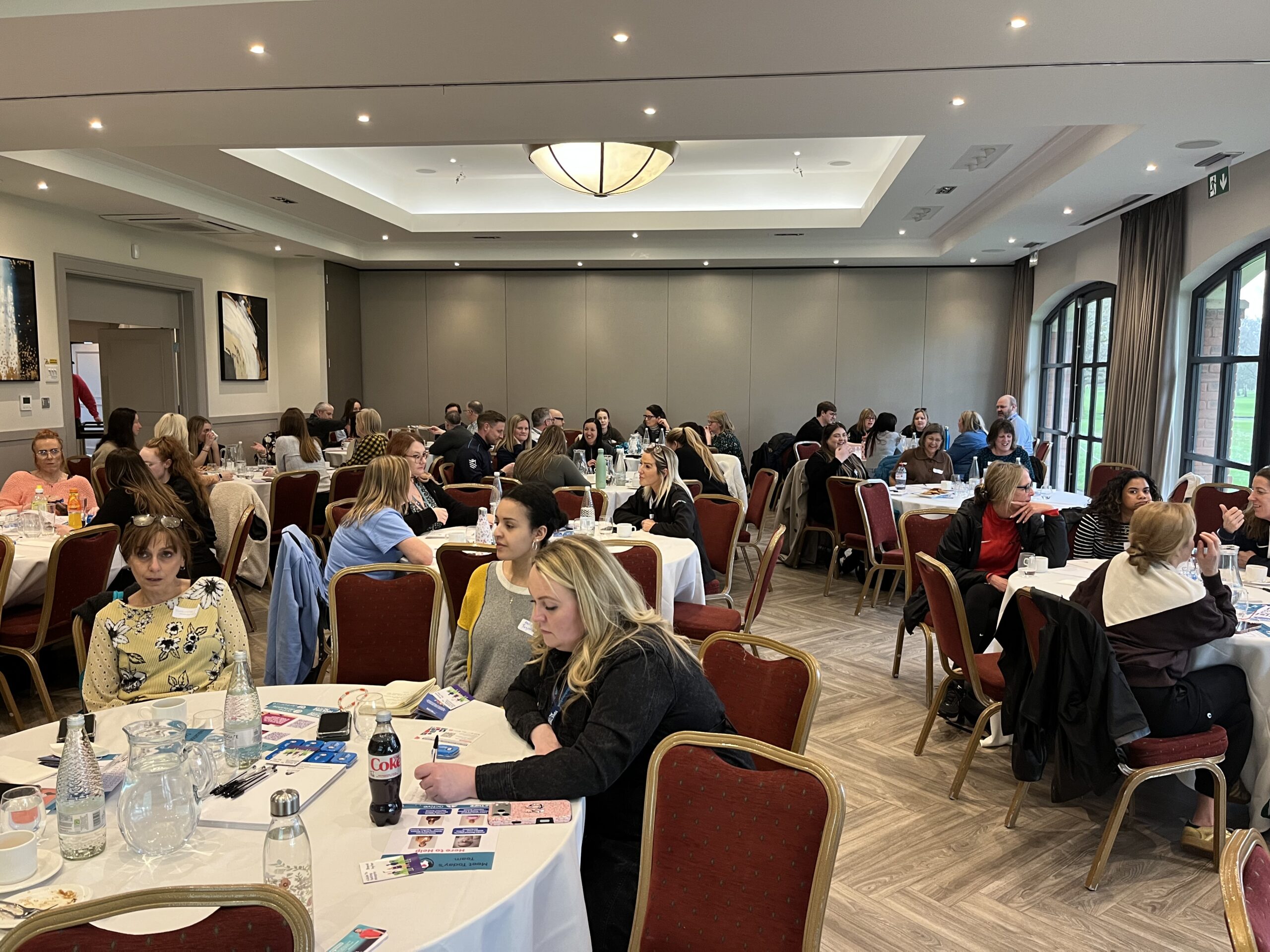 Image shows a large conference room with people sat around round tables in discussion.